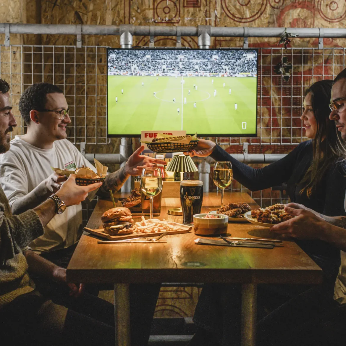 Group of friends sharing burgers, chicken wings, fries and Guinness at Belushi's sports bar with a dedicated screen showing the World Cup live behind them