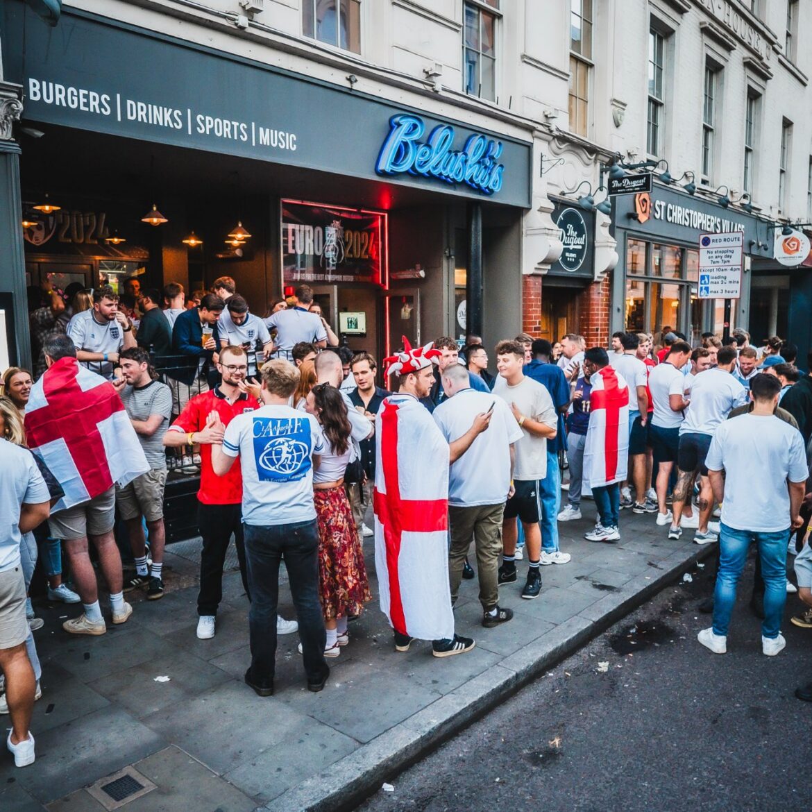 Long queue of England football fans outside Belushi's London Bridge sports bar waiting to watch a World Cup 2026 match, with St George's Cross flags and the Belushi's neon sign above the entrance