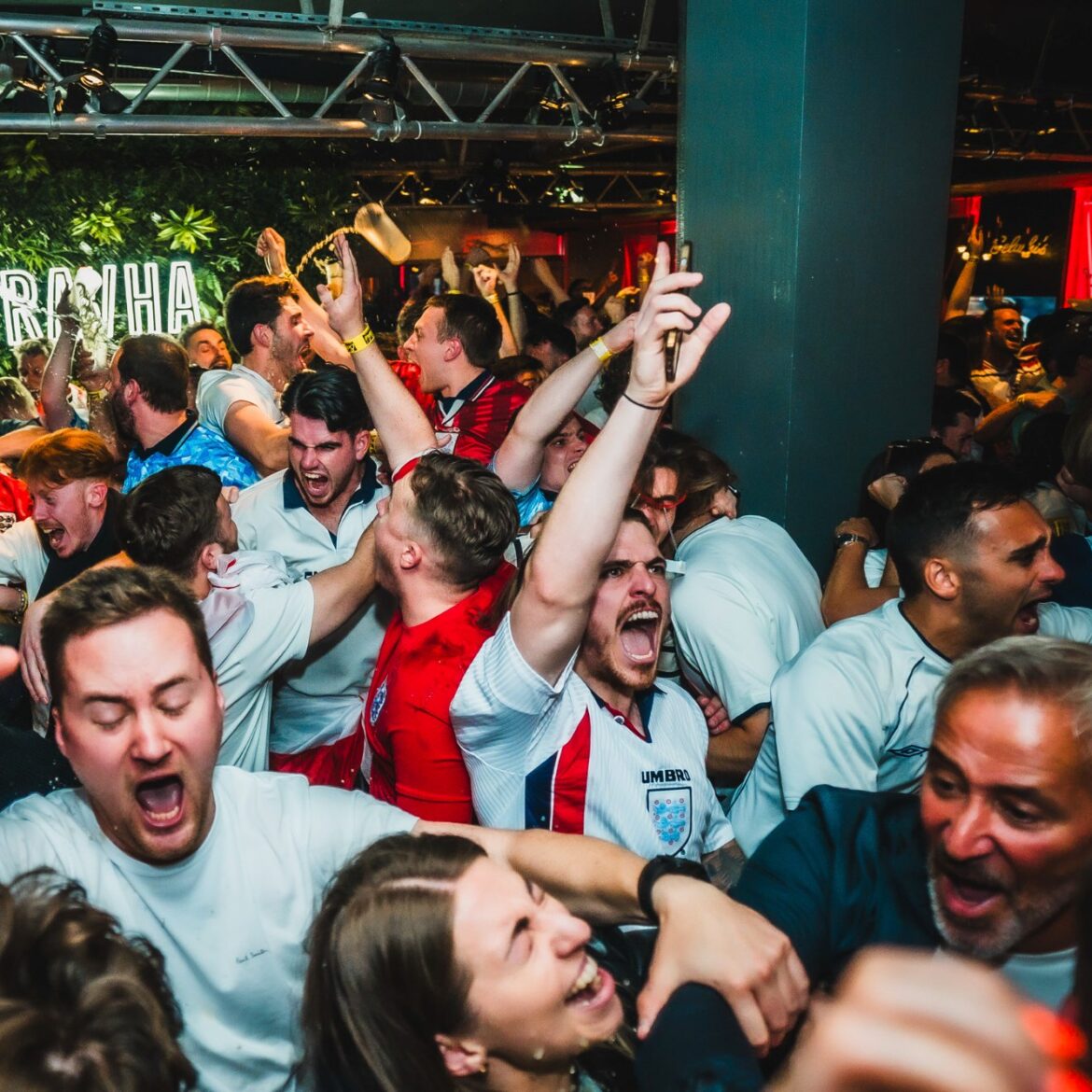 Packed crowd of England football fans erupting in celebration at Belushi's Shepherd's Bush after a World Cup goal, with confetti in the air and the Pravha bar area in the background