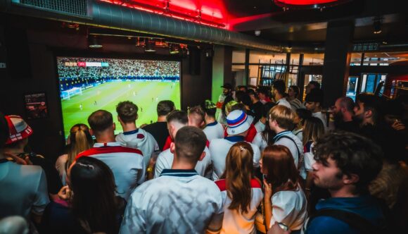 Crowd of football fans watching a World Cup 2026 match live on a large projector screen at Belushi's Greenwich, with red overhead lighting and a packed standing crowd