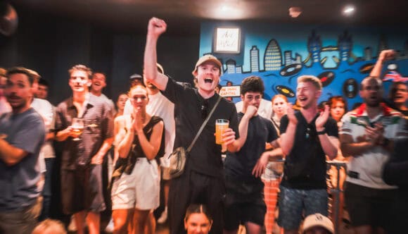 Crowd of people at a party cheering, central man raises a fist and holds a beer with a bright mural behind them.